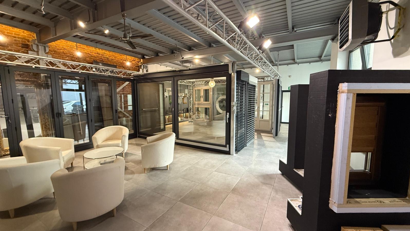 Salon ekspozycyjny z oknami, drzwiami i roletami. Widoczne różne modele i systemy, krzesła dla klientów i oświetlenie sufitowe.