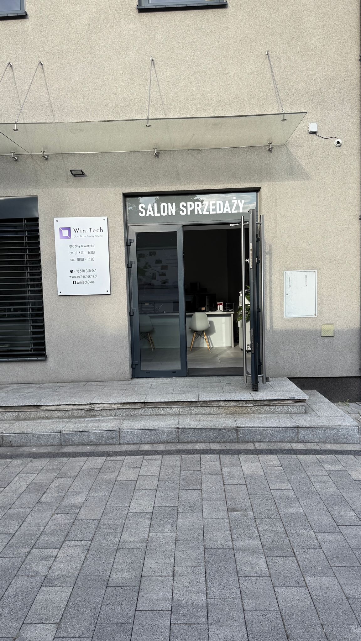 Wejście do salonu sprzedaży Win-Tech z otwartymi szklanymi drzwiami, widoczna tabliczka z godzinami otwarcia i logo firmy. Nad wejściem szklany daszek.