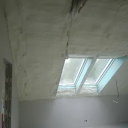 Poddasze po natrysku pianką PUR, widoczne okno dachowe i zwisający kabel z żarówką.