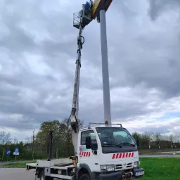 Podnośnik koszowy na parkingu, pracownik w koszu przy neonie Biedronki. Biała ciężarówka z wysięgnikiem na tle pochmurnego nieba.