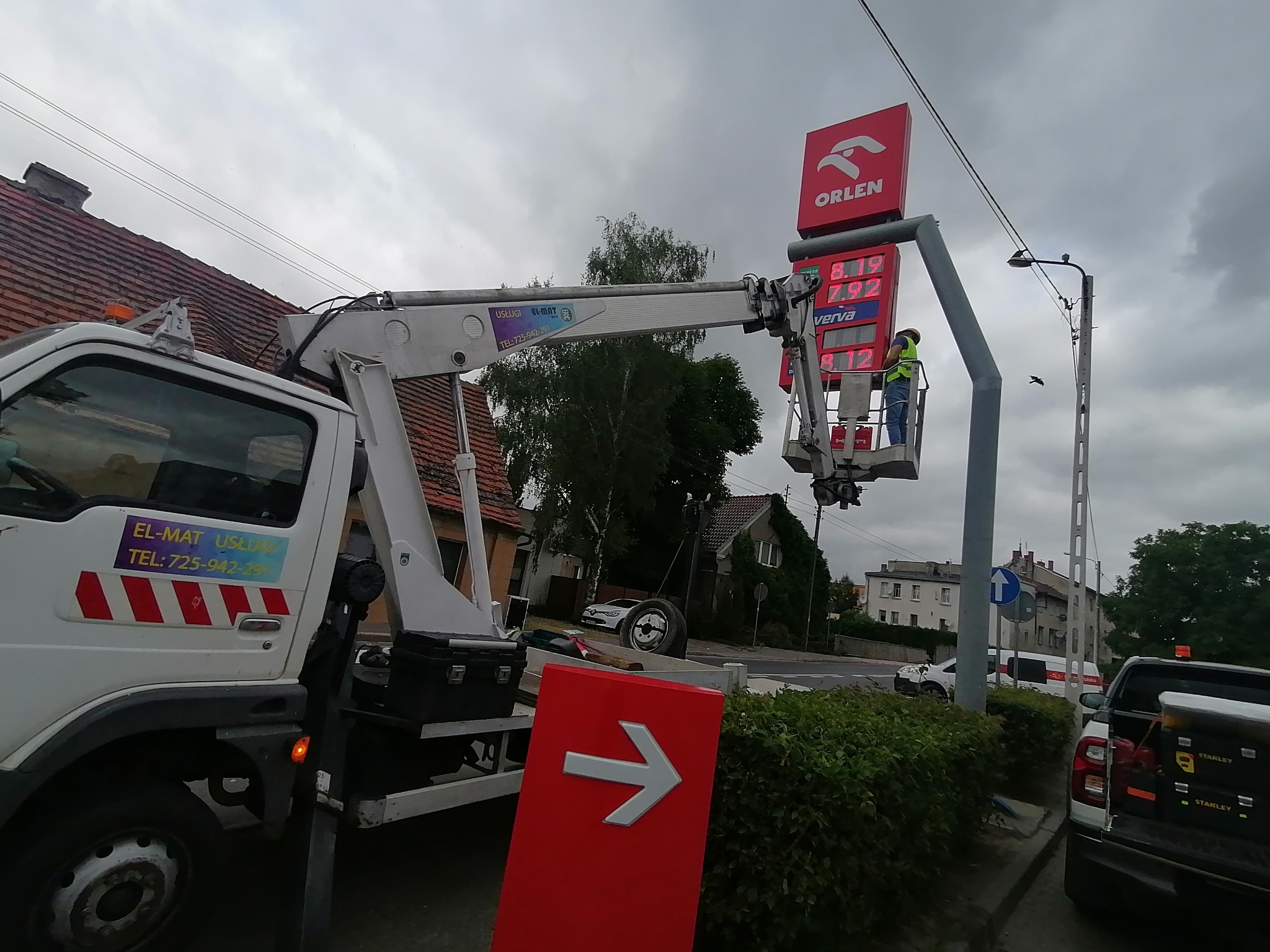 Pracownik w koszu podnośnika przy pylonie stacji benzynowej Orlen, prawdopodobnie wymiana lub naprawa oświetlenia, widoczny samochód z podnośnikiem z naklejką firmy EL-MAT.
