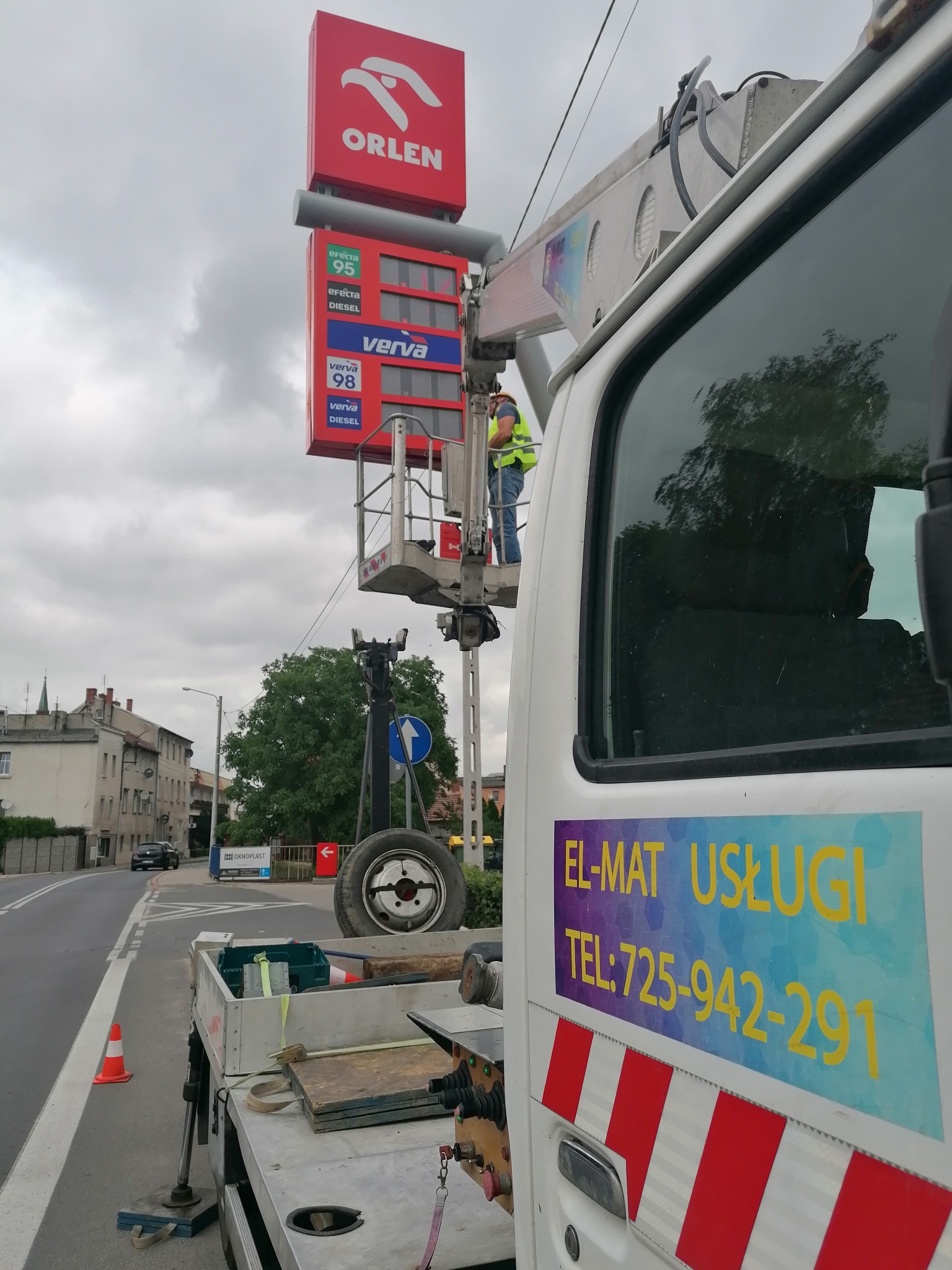 Pracownik w koszu podnośnika konserwuje pylon cenowy stacji paliw Orlen, widoczne elementy konstrukcyjne podnośnika oraz reklama firmy EL-MAT USŁUGI na pojeździe.