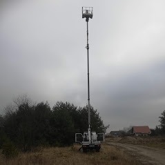 Podnośnik koszowy na pojeździe, wysunięty na maksymalną wysokość, na tle zalesionego terenu i pochmurnego nieba.