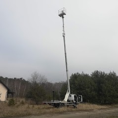 Wysięgnik koszowy na pojeździe ciężarowym, rozłożony na maksymalną wysokość, widoczny na tle drzew i pochmurnego nieba, z domem jednorodzinnym w lewym dolnym rogu.
