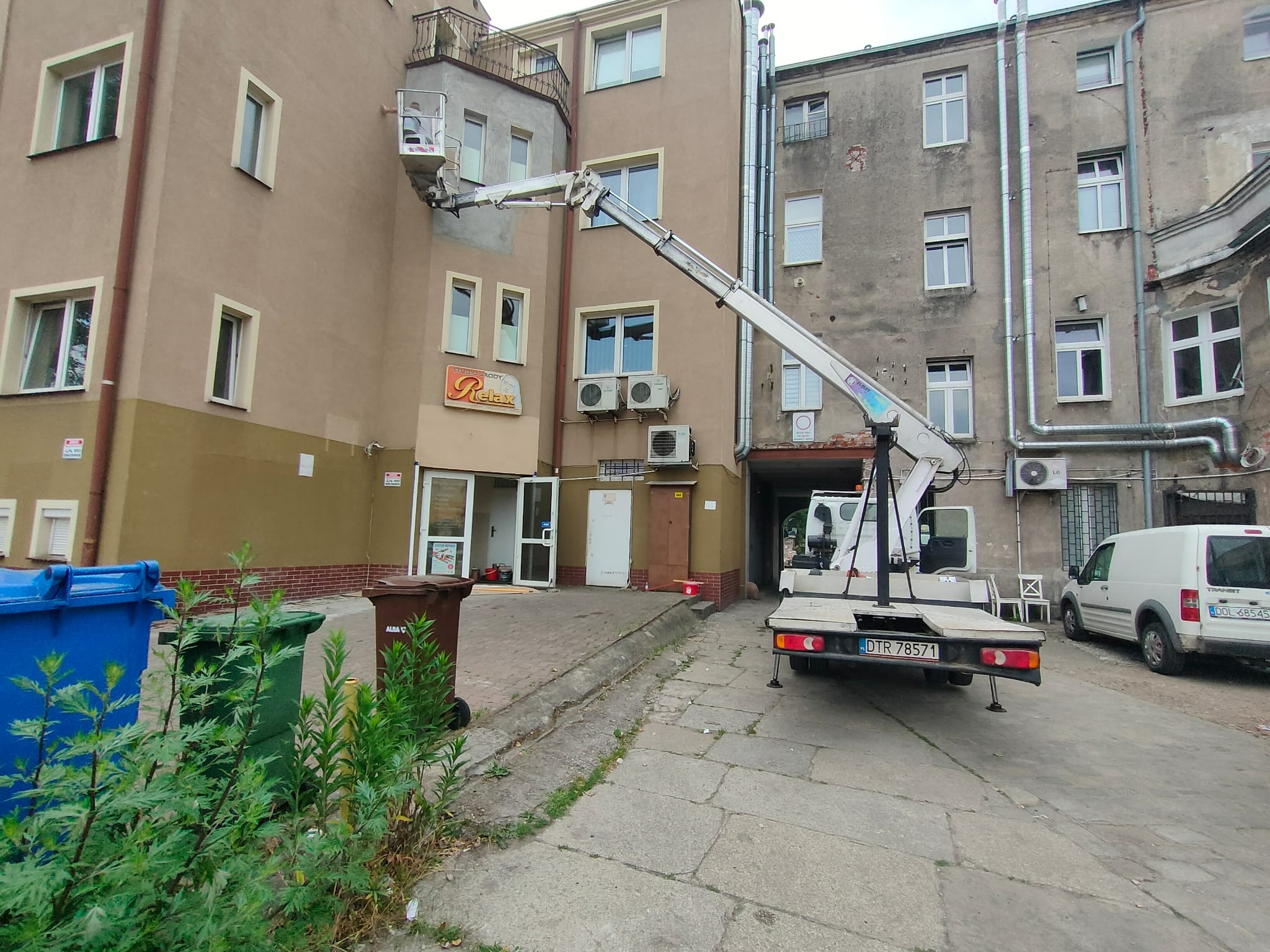 Podnośnik koszowy przed budynkiem z szyldem 'Relax'. Widoczne kosze na śmieci i zaparkowany van. Prace na wysokości, dostęp do trudno dostępnych miejsc.