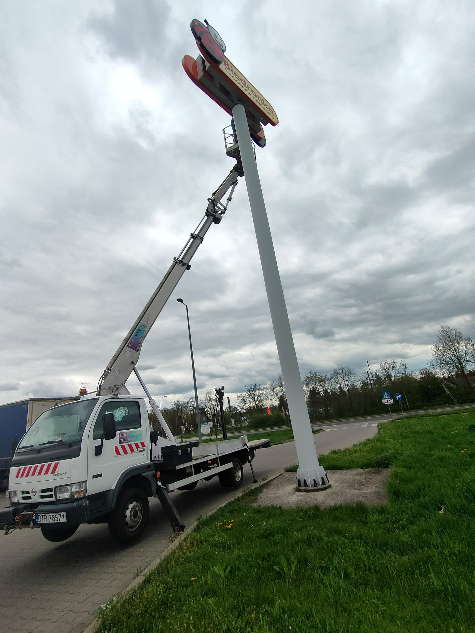 Podnośnik koszowy przy montażu dużego szyldu reklamowego Biedronki. Widoczny biały samochód z podnośnikiem i wysięgnikiem. Pochmurne niebo w tle.
