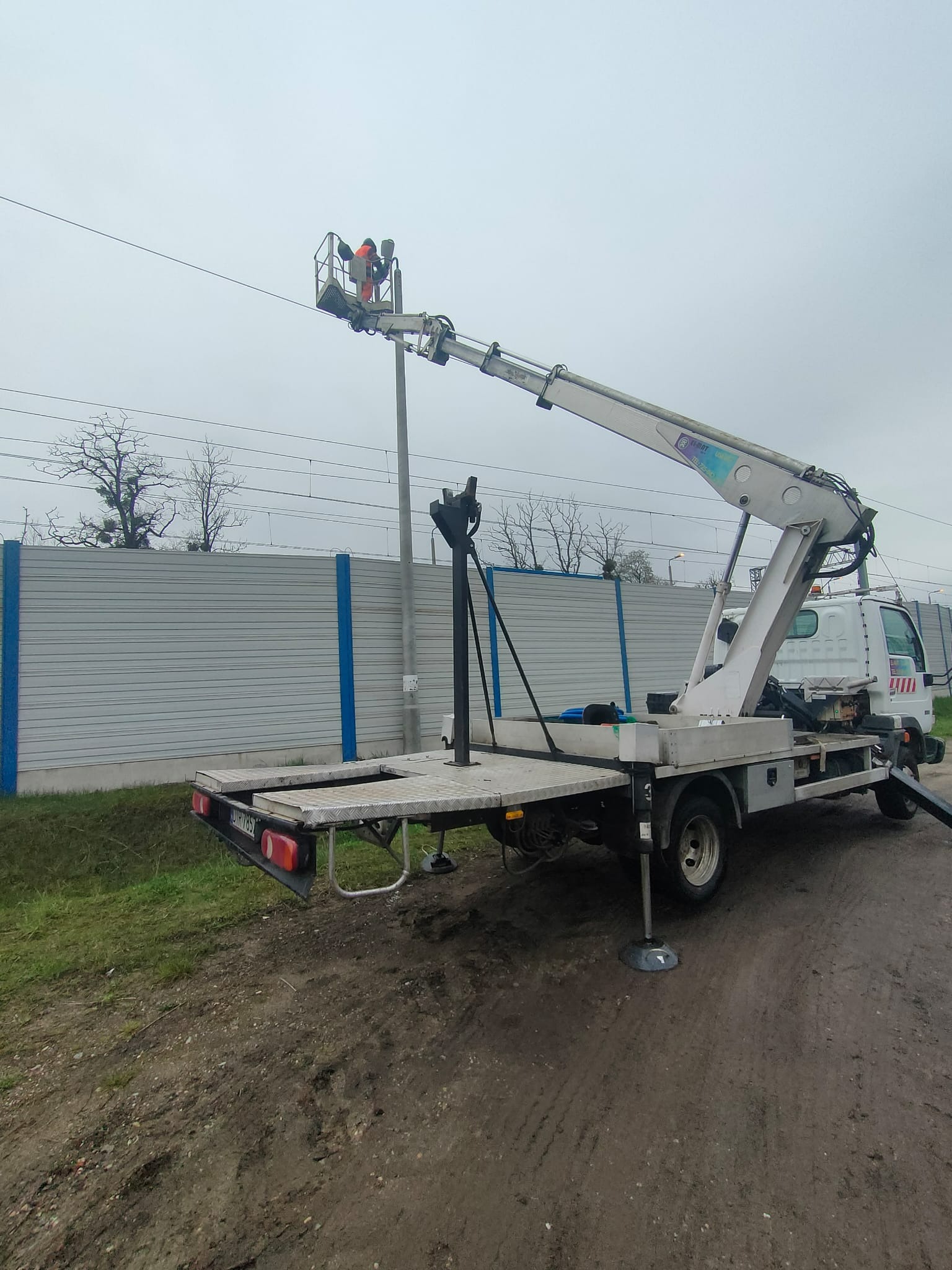 Podnośnik koszowy ustawiony na nierównym terenie, pracownik w pomarańczowym kombinezonie w koszu, szara bariera akustyczna w tle.