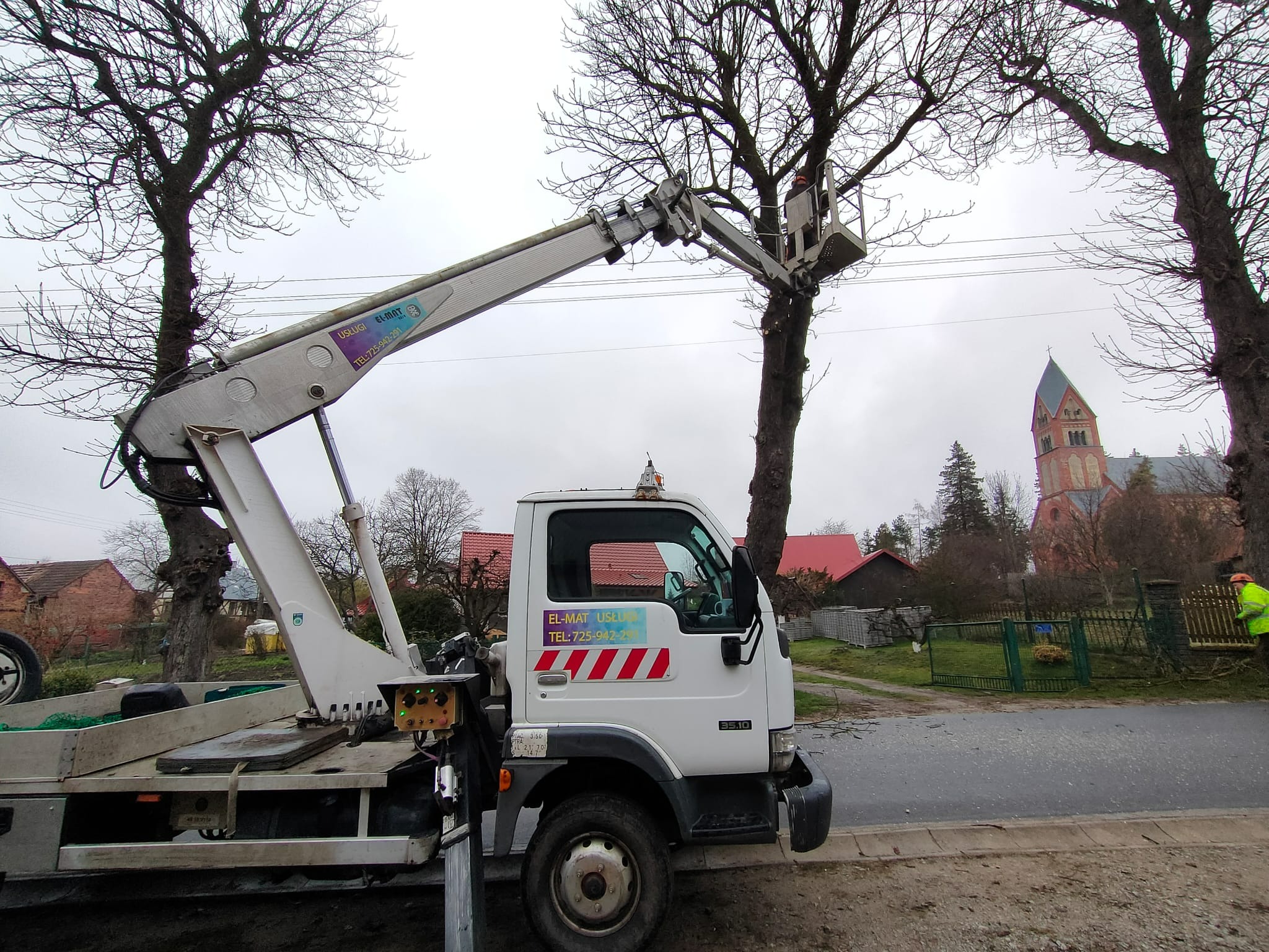 Pracownik w koszu podnośnika przycina gałęzie drzewa. W tle widoczny kościół i zabudowa wiejska. Sprzęt z logo firmy EL-MAT USŁUGI.