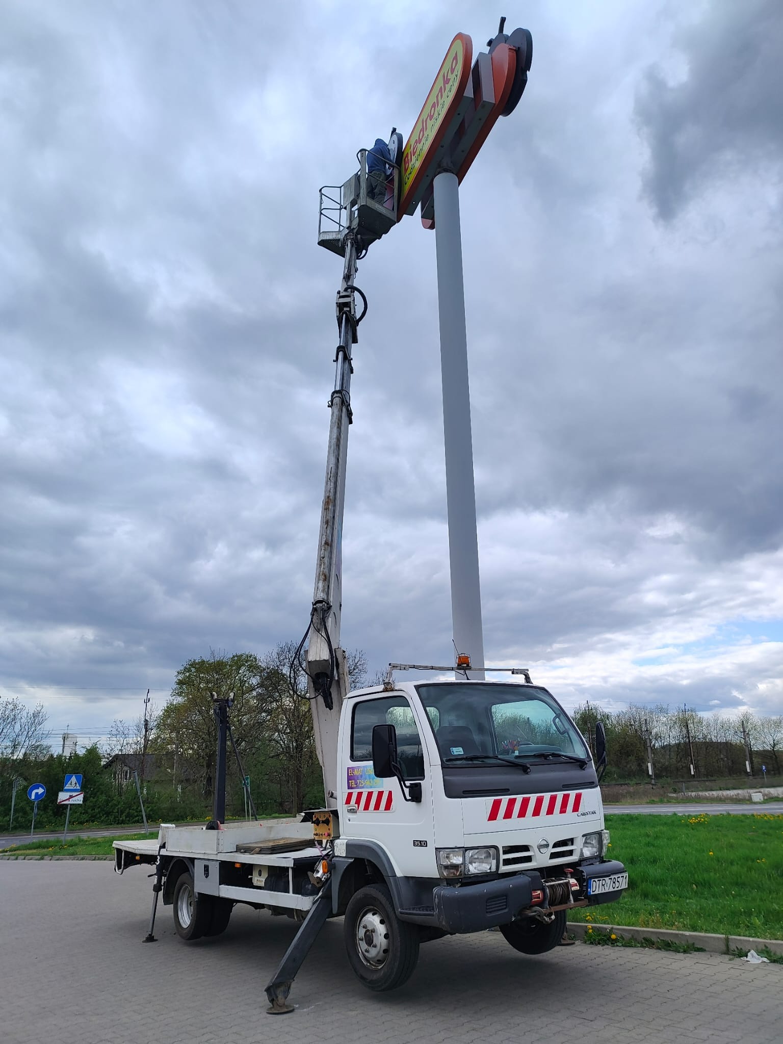 Podnośnik koszowy na parkingu, pracownik w koszu przy neonie Biedronki. Biała ciężarówka z wysięgnikiem na tle pochmurnego nieba.