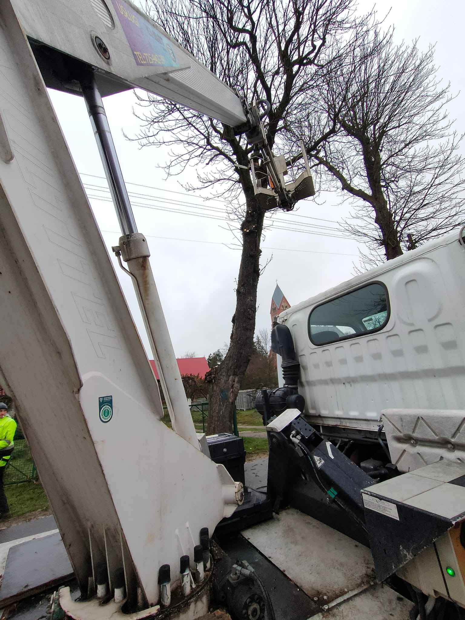Biały podnośnik koszowy w trakcie wycinki gałęzi drzewa w pochmurny dzień. W tle widoczny budynek z czerwoną dachówką i wieżą kościoła.