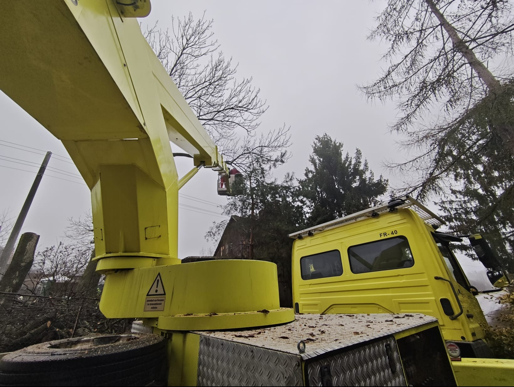 Żółty podnośnik koszowy z wysięgnikiem i koszem na tle drzew i pochmurnego nieba, widoczne ścięte gałęzie. Prace w toku.