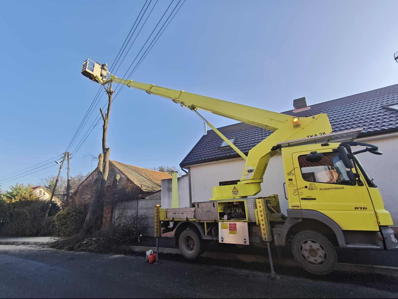 Żółty podnośnik koszowy TKA 28 przy ogołoconym drzewie, z pracownikiem w koszu na tle błękitnego nieba i zabudowań. Ścięte gałęzie leżą na ziemi.