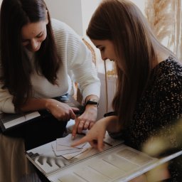 About The Form Weronika Sypuła - Dwie kobiety przeglądają próbki materiałów i kolorów z katalogu, siedząc na tle beżowej ściany i dekoracyjnego makramy.