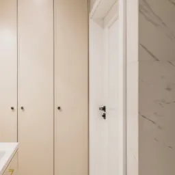 Nowoczesna łazienka z beżowymi szafkami, białymi drzwiami z czarną klamką i marmurowymi płytkami na ścianie, uzupełniona geometryczną mozaiką podłogową.