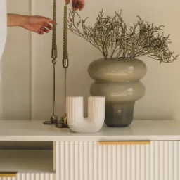 Kobieta ustawia oliwkową, skręconą świecę na złotym świeczniku obok wazonu z suszonymi kwiatami i dekoracyjnej ceramicznej rzeźby na białej komodzie z ryflowanymi drzwiami i złotymi uchwytami.