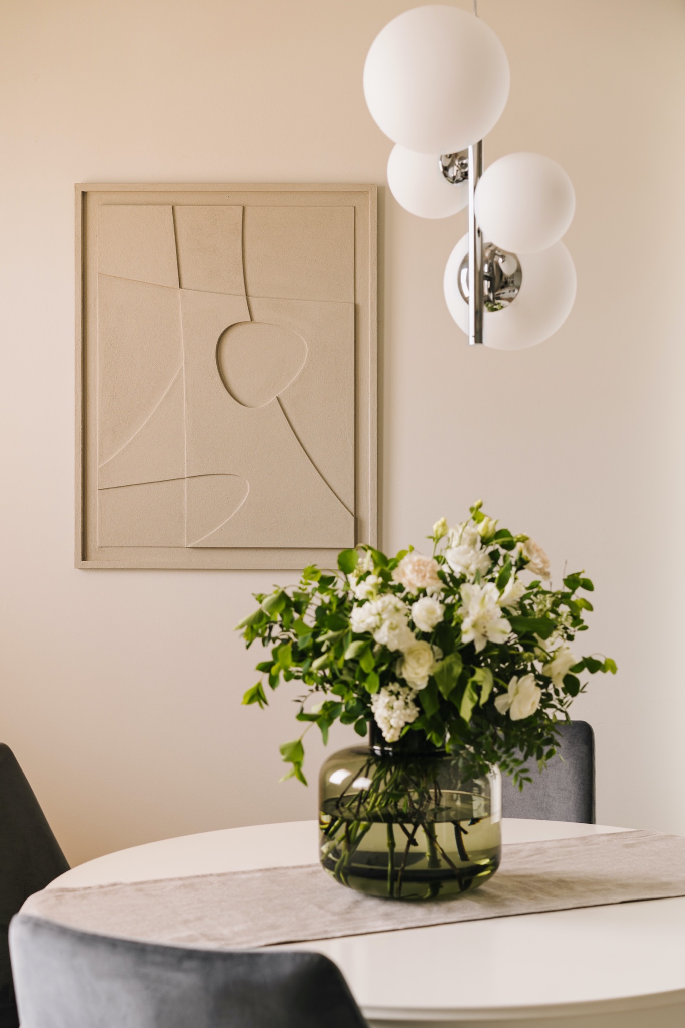 Beżowe, minimalistyczne wnętrze: abstrakcyjna płaskorzeźba na ścianie, okrągły stół z szarą bieżnikiem, bukiet białych kwiatów w szklanym wazonie i lampa sufitowa z czterema mlecznymi kulami.