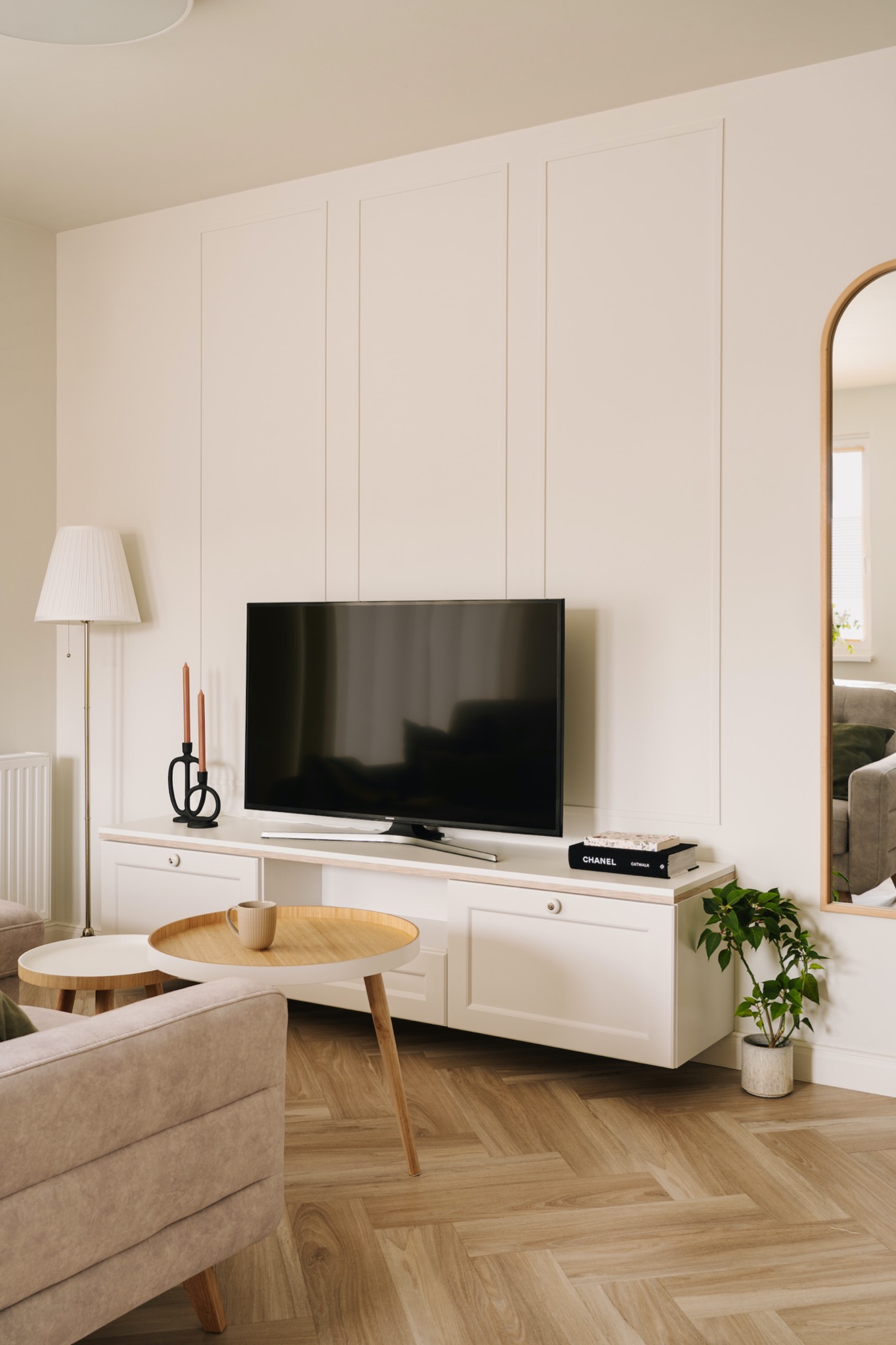 Jasne wnętrze salonu z parkietem w jodełkę, kremową ścianą z panelami, białym regałem RTV, telewizorem, lampą podłogową, okrągłymi stolikami kawowymi i lustrem w złotej ramie.