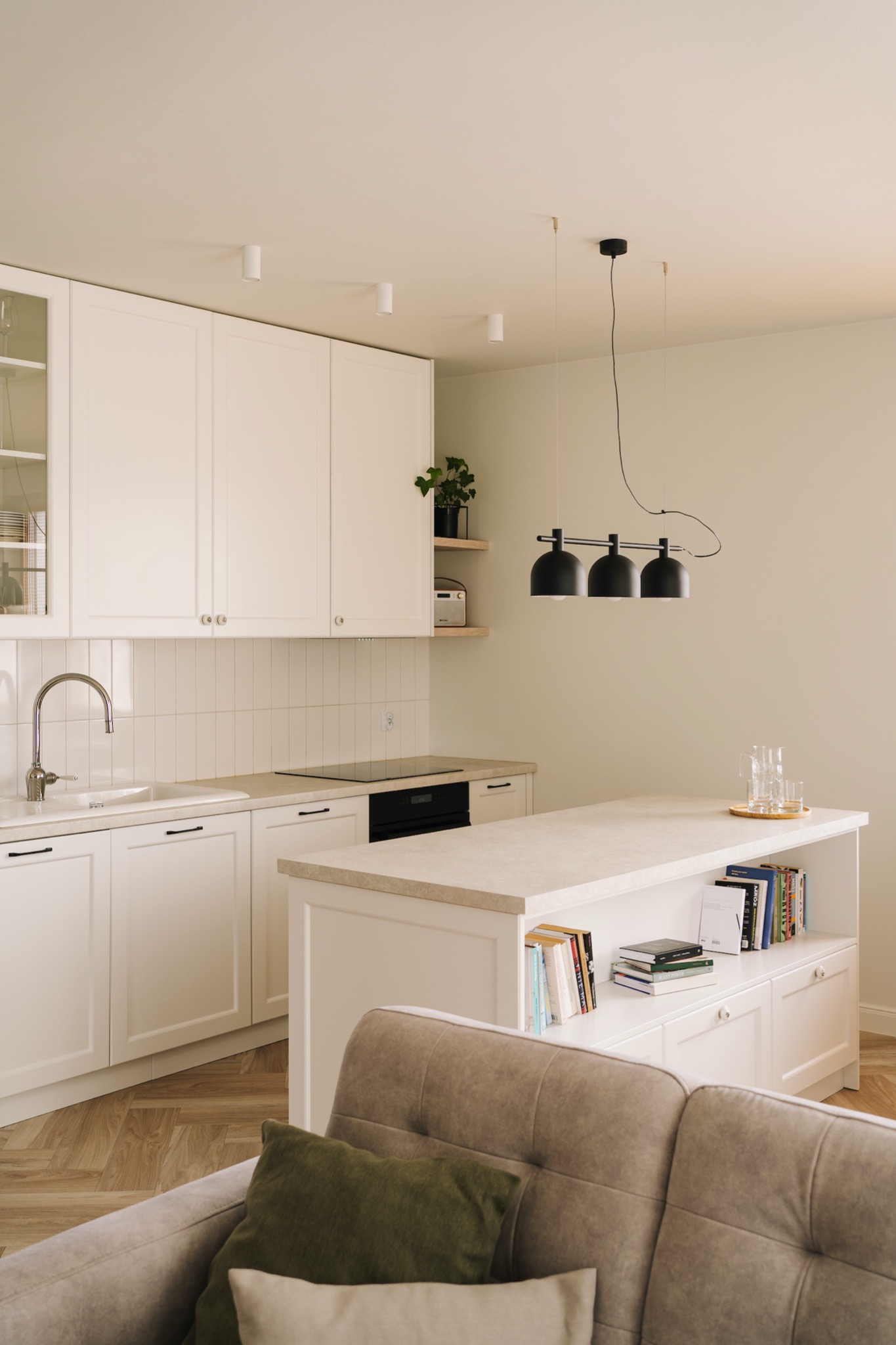 Jasna kuchnia z wyspą, białymi szafkami, drewnianą podłogą w jodełkę i czarną lampą wiszącą nad wyspą. Widoczny fragment sofy z poduszkami.
