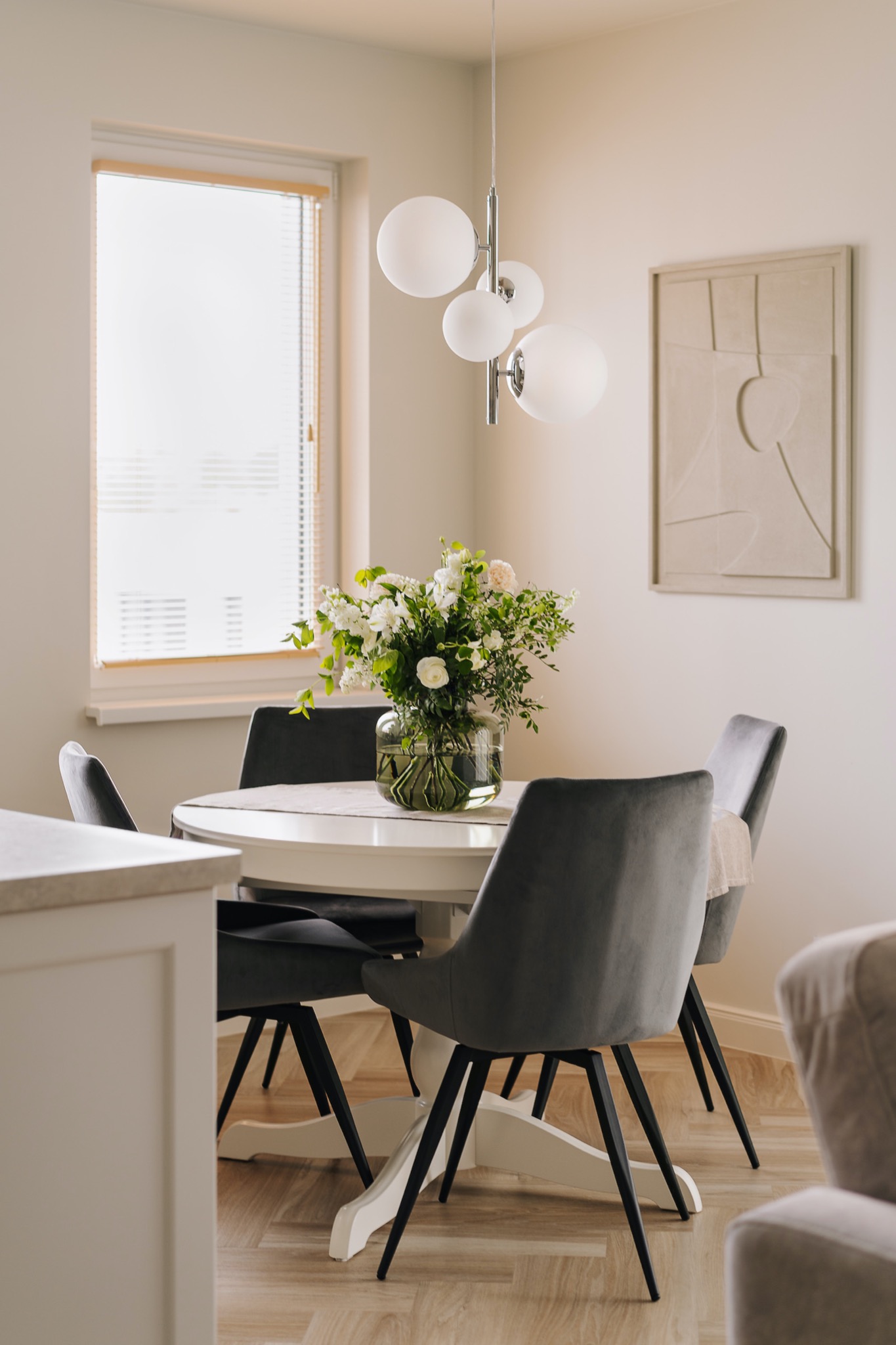 Jasne wnętrze jadalni z okrągłym białym stołem, szarymi krzesłami na czarnych nogach, bukietem białych kwiatów w szklanym wazonie, wiszącą lampą z białymi kulami i abstrakcyjnym obrazem na ścianie.