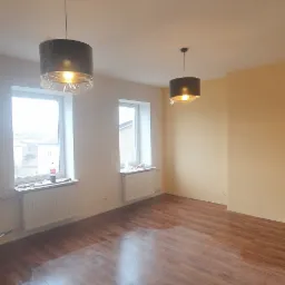 Pusty pokój po malowaniu ścian na jasny beż, z nową, ciemną podłogą panelową i dwoma wiszącymi lampami z abażurami w folii ochronnej.