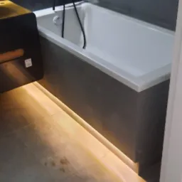 Nowoczesna biała wanna z czarną armaturą, podświetlona listwą LED u dołu obudowy, na tle szarych płytek ściennych i podłogowych.