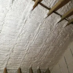 Poddasze z drewnianą konstrukcją wypełnione białą pianką PUR, widok z dołu, ukazujący strukturę ocieplenia i szczelne wypełnienie przestrzeni.