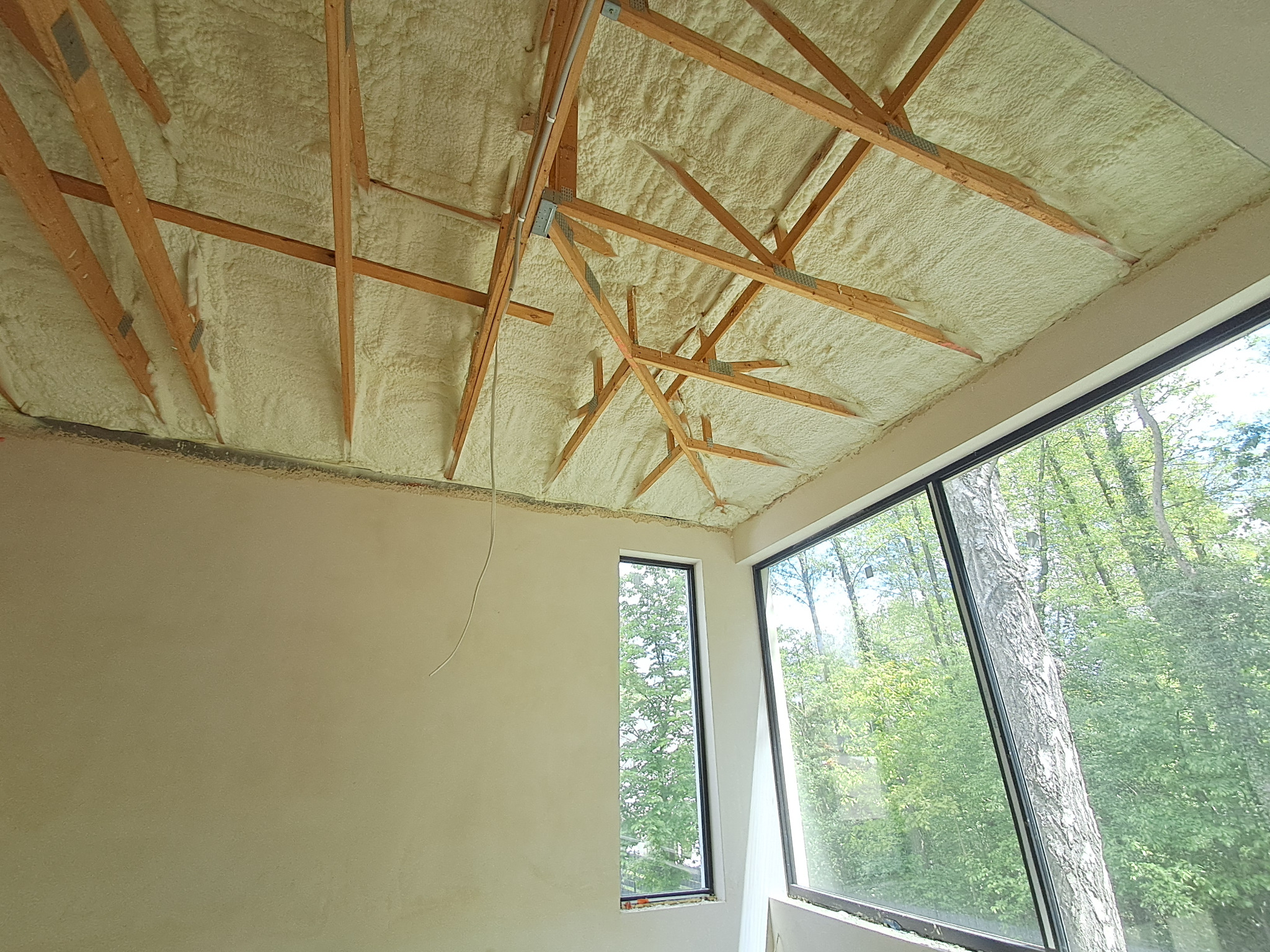 Poddasze po ociepleniu pianką PUR, widoczne drewniane krokwie i warstwa izolacji. Okna z widokiem na zieleń. Detal z bliska,  jasne oświetlenie.