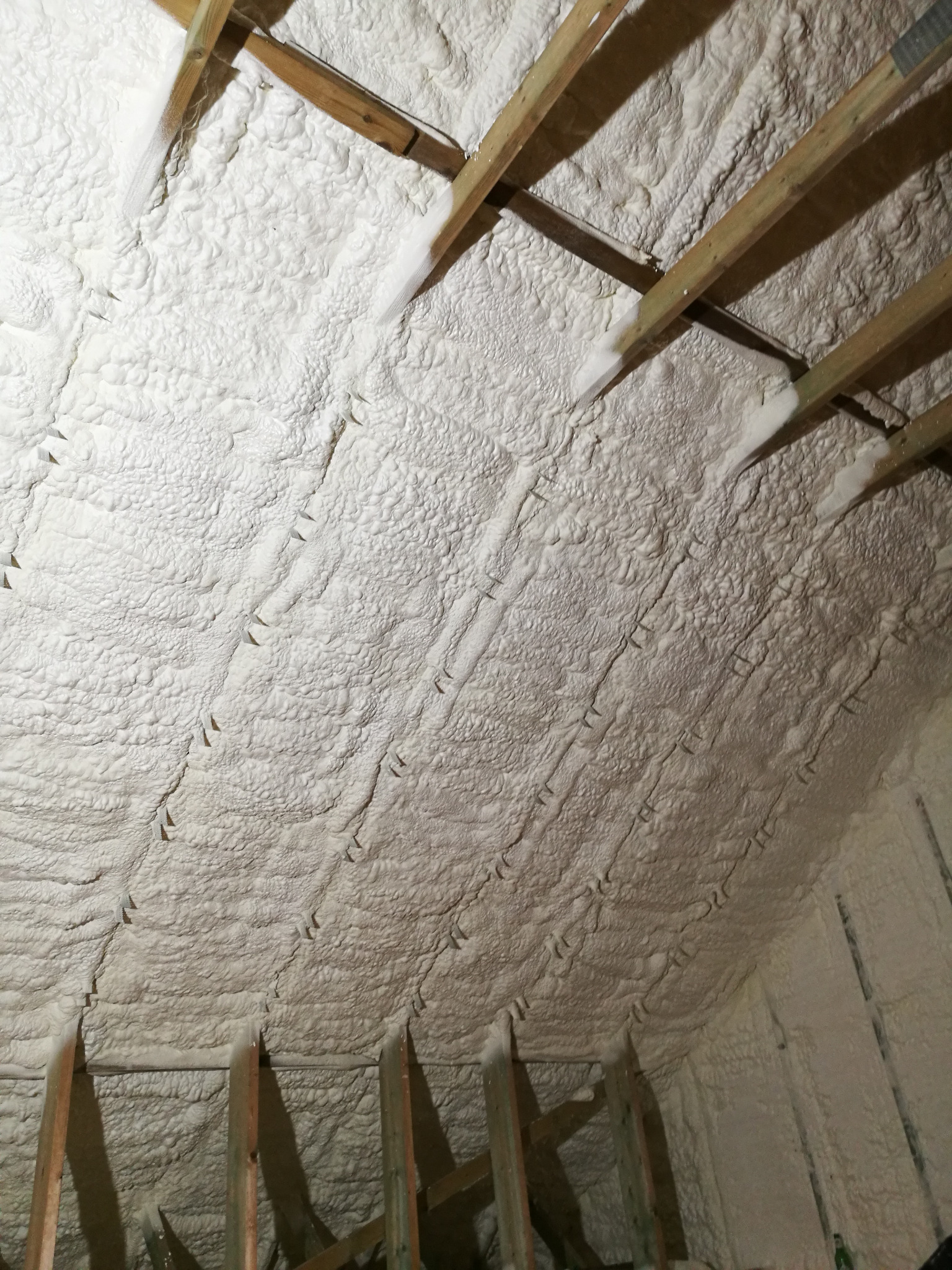 Poddasze z drewnianą konstrukcją wypełnione białą pianką PUR, widok z dołu, ukazujący strukturę ocieplenia i szczelne wypełnienie przestrzeni.