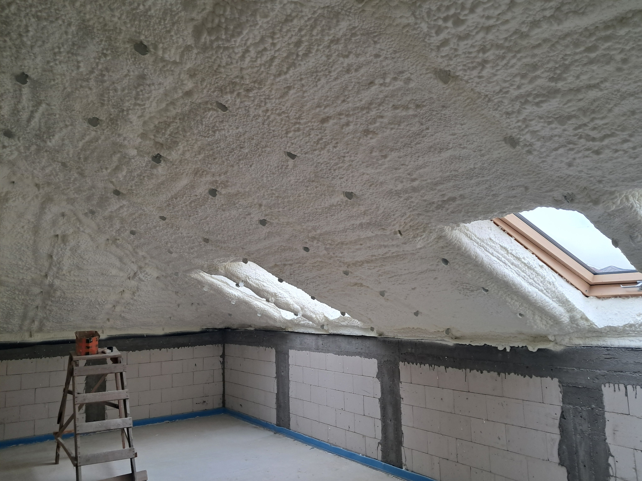 Poddasze ocieplone pianką PUR, widoczne otwory wentylacyjne i okno dachowe. Ściany z bloczków, drabina z farbą w rogu.