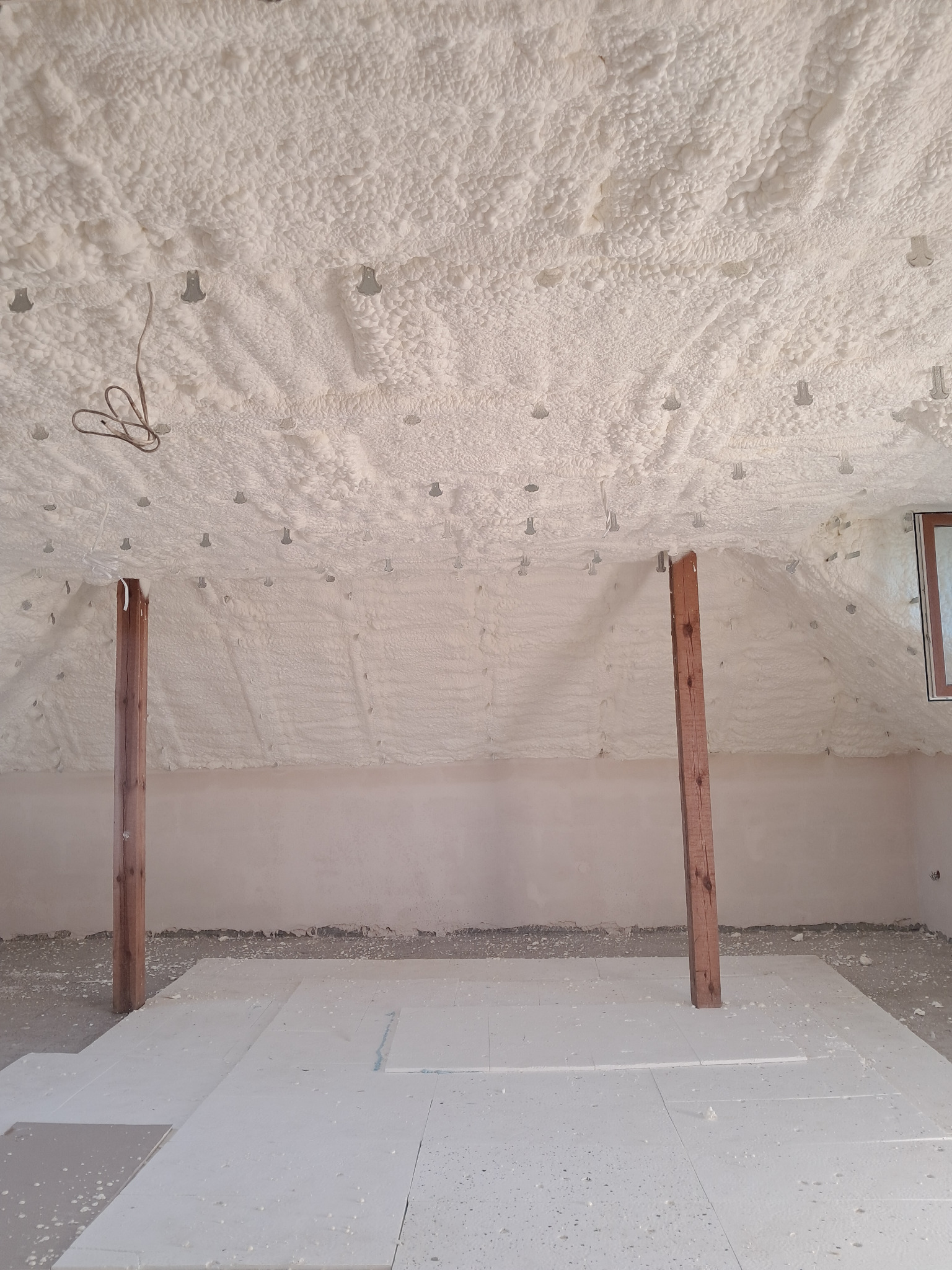 Poddasze po ociepleniu pianką PUR, widoczne drewniane słupy, biała pianka na skosach i suficie, oraz płyty izolacyjne na podłodze.