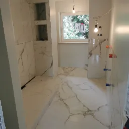 Wnętrze łazienki w trakcie wykańczania, widoczne białe płytki imitujące marmur na podłodze i ścianach, wnęki w ścianie oraz wystające kable i puszki elektryczne.