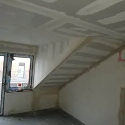 Poddasze w trakcie prac wykończeniowych: widoczne szpachlowane łączenia płyt karton-gips na skośnym suficie i ścianach, z instalacją elektryczną zabezpieczoną r&oacute;żową pianką montażową.