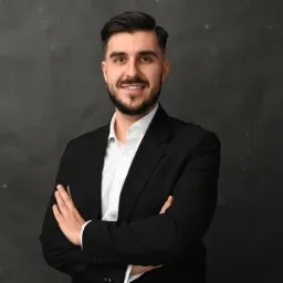 Portret uśmiechniętego mężczyzny z brodą w czarnym garniturze i białej koszuli, stojącego na tle ciemnoszarej ściany z założonymi rękami.