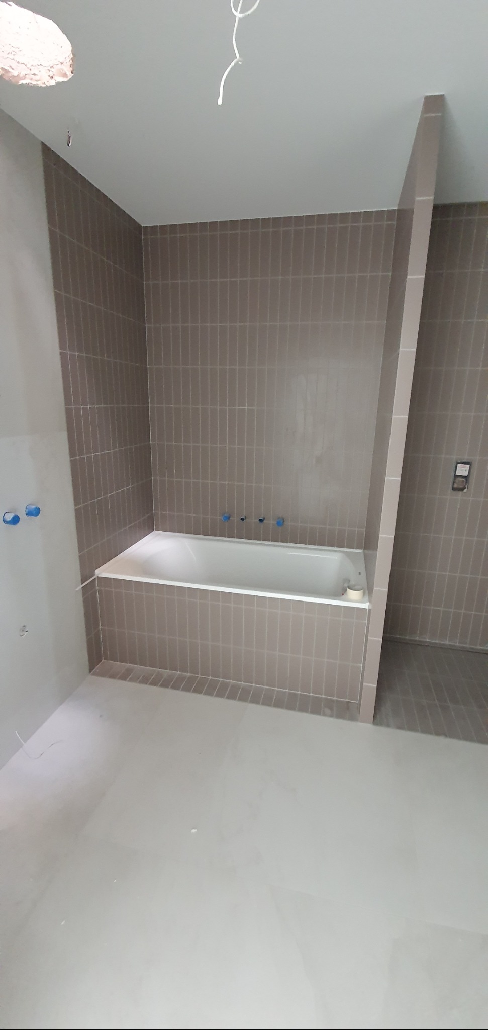 Wnętrze łazienki w trakcie prac wykończeniowych, widoczna wanna obudowana płytkami w odcieniach szarości, ściany wyłożone pionowymi płytkami, niezagruntowana podłoga i wystające kable ze stropu.