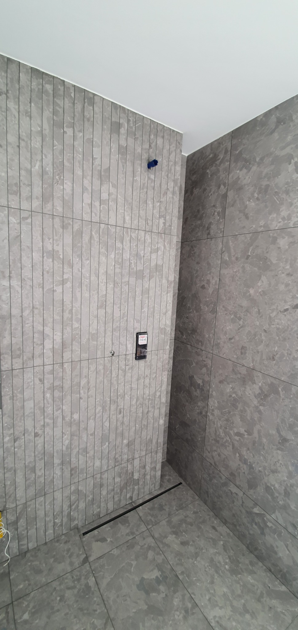 Szare płytki imitujące marmur na ścianach i podłodze w łazience, widoczny odpływ liniowy i wystające kable.