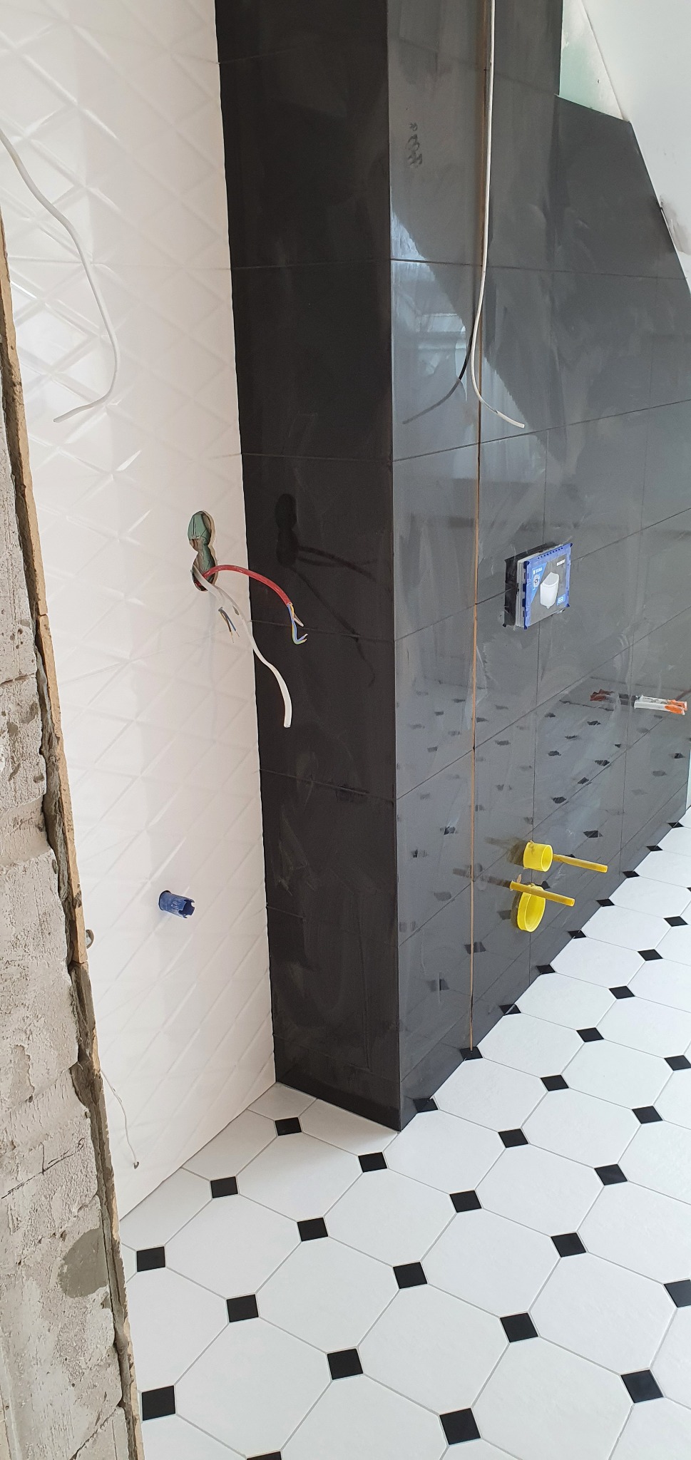 Częściowo wykończona łazienka: białe płytki ścienne z trójwymiarowym wzorem, czarne płytki na pionowym elemencie oraz biało-czarna podłoga w romby, widoczne przewody elektryczne i elementy...