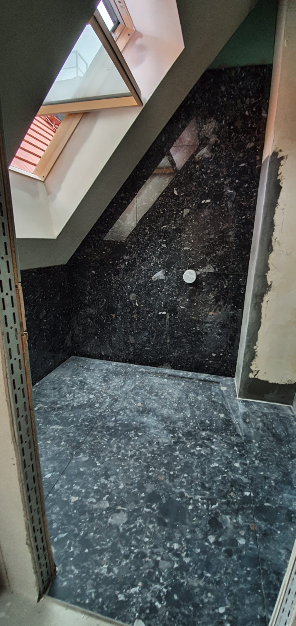 Łazienka w trakcie remontu na poddaszu, wykończona czarnym lastryko na ścianach i podłodze, widoczne okno dachowe i niedokończone obrzeża ścian.