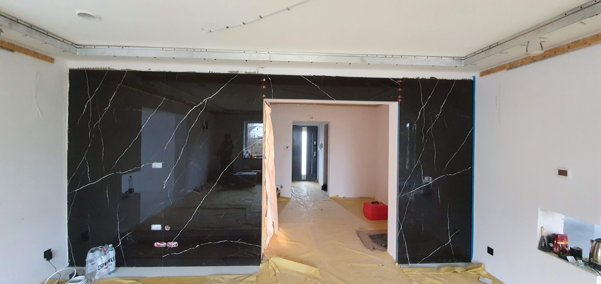 Montaż czarnych płyt imitujących marmur na ścianie w trakcie remontu, widoczne profile sufitowe, folia ochronna na podłodze i otwór drzwiowy do kolejnego pomieszczenia.
