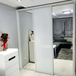 Biała szafa przesuwna z lustrem i białymi frontami na tle jasnego wnętrza pokoju z komodą, kwiatami i widocznym fragmentem sofy.