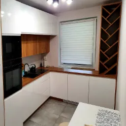 Nowoczesna kuchnia z białymi szafkami i blatem z jasnego drewna, czarny zlew i bateria, drewniany regał na wino, koszyk z owocami na stole.