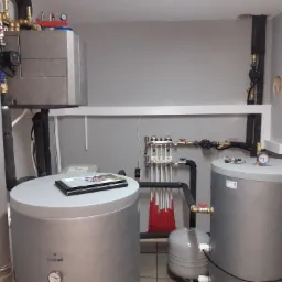 Instalacja grzewcza w kotłowni: dwa szare zbiorniki Galmet z manometrami, rozdzielacz z rurami do ogrzewania podłogowego, obiegowa pompa ciepła, szara ściana i czarne rury izolowane.