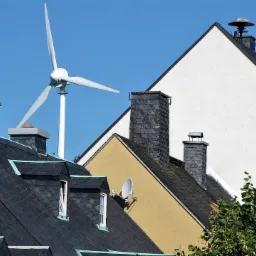 Turbina wiatrowa na tle nieba i dachów budynków o różnej architekturze i kolorystyce, widoczna antena satelitarna.