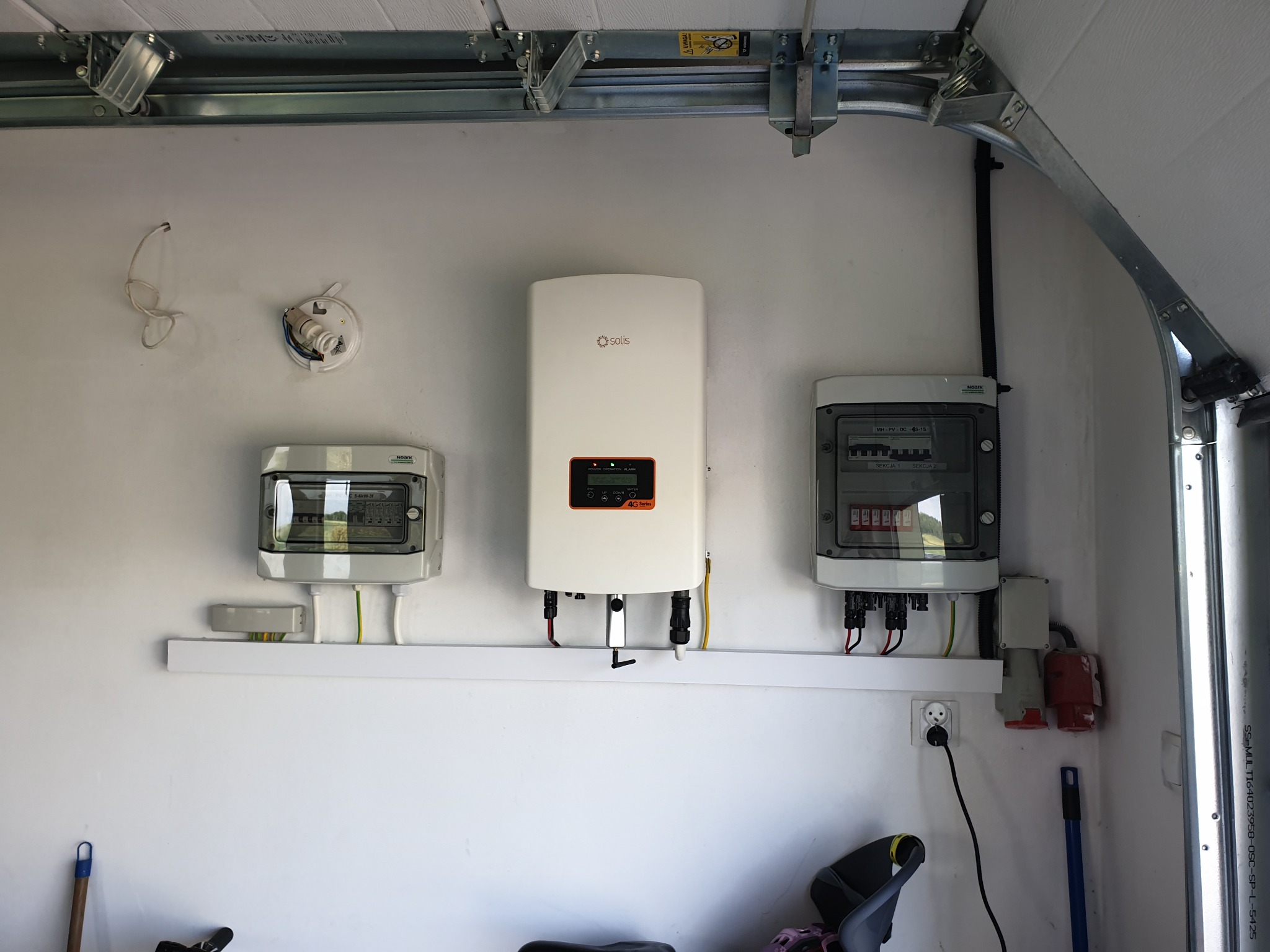 Biały falownik z wyświetlaczem, dwie szare skrzynki elektryczne z widocznymi bezpiecznikami, zamontowane na ścianie garażu nad białą półką z przewodami, widoczne gniazdko elektryczne i fragment...