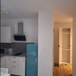 Wnętrze kuchni developerskiej z turkusową lodówką Amica, szarymi płytkami nad blatem i widokiem na otwartą szafę w przedpokoju.