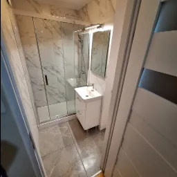 Nowoczesna łazienka po remoncie: kabina prysznicowa z przesuwnymi drzwiami, umywalka z szafką w kolorze białym, płytki imitujące marmur na ścianach i podłodze, oświetlenie LED nad lustrem.