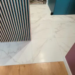 Białe płytki gresowe imitujące marmur, ułożone w kuchni, zlicowane z drewnianą podłogą w salonie. Widoczna dekoracyjna ścianka z drewnianych listewek z turkusowym wypełnieniem.