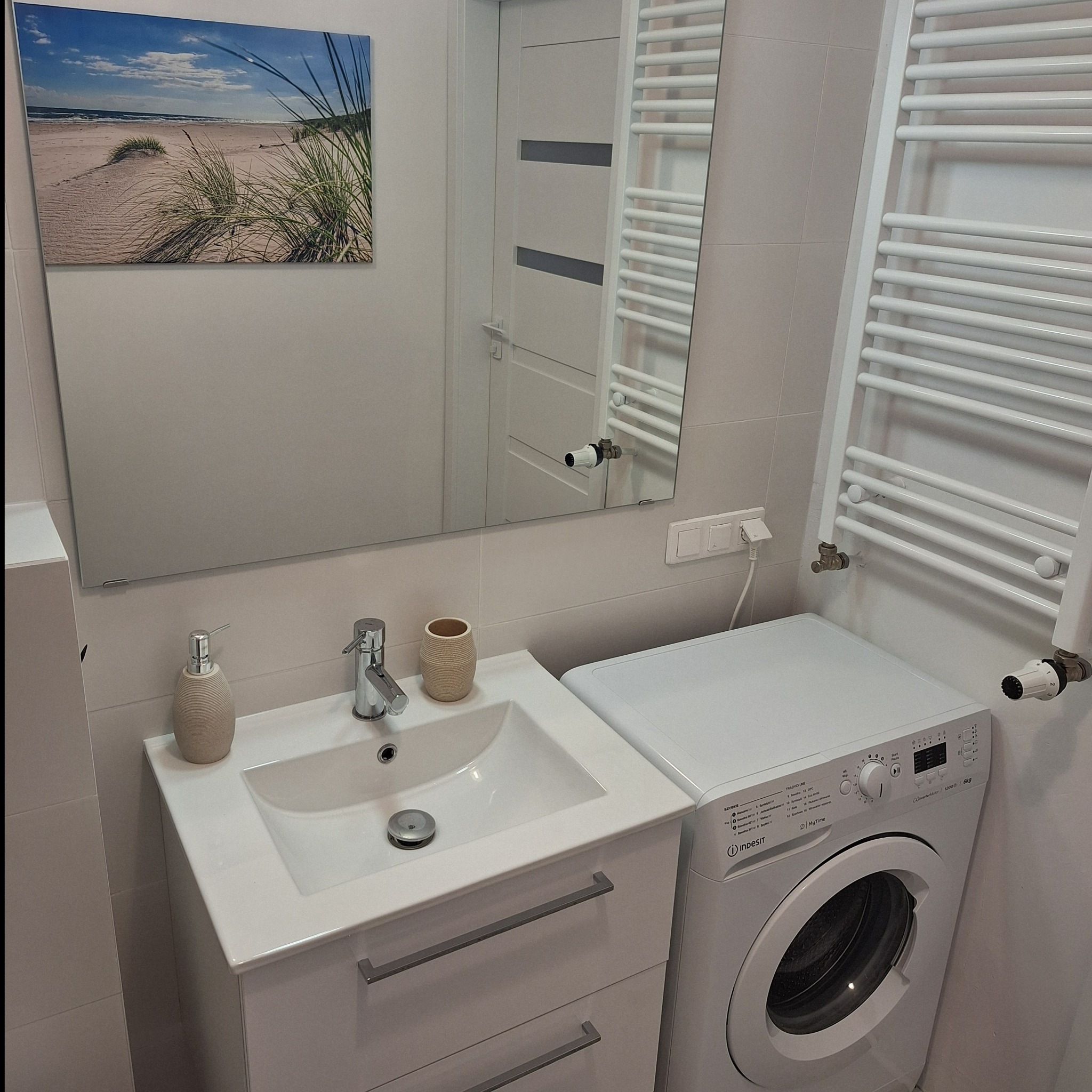 Nowoczesna łazienka w stanie developerskim: biała umywalka z szafką, pralka Indesit, grzejnik drabinkowy i lustro z widocznym obrazem plaży.