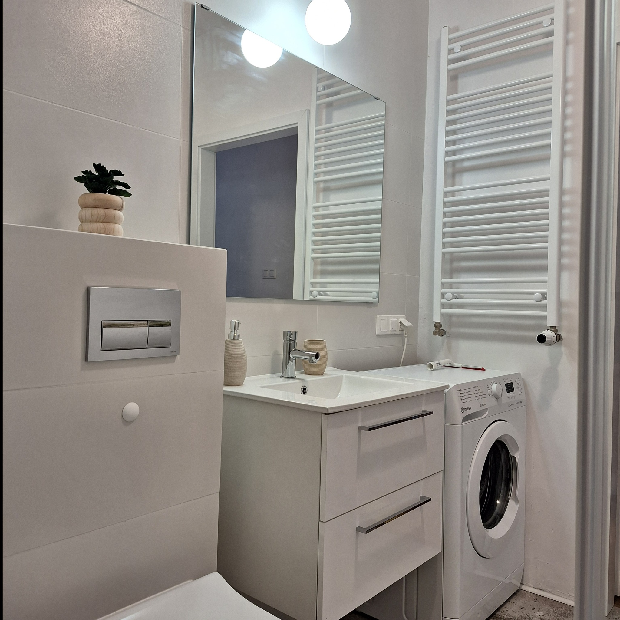 Nowoczesna łazienka w stanie deweloperskim z białymi płytkami, umywalką z szafką, lustrem z oświetleniem, grzejnikiem drabinkowym i pralką Indesit.