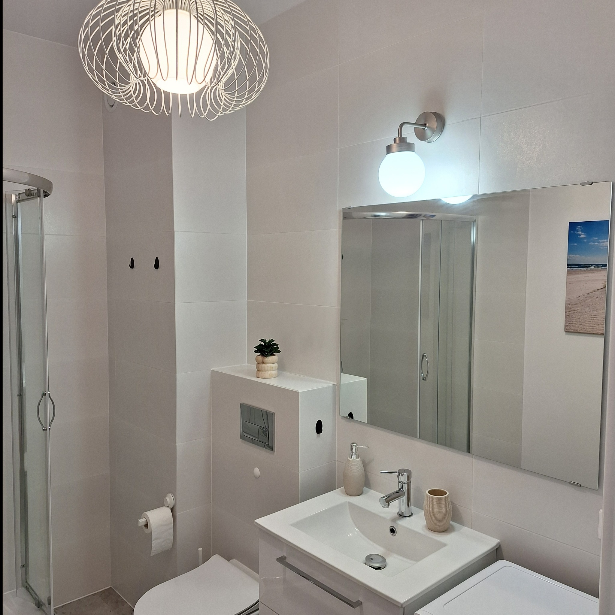 Jasna łazienka w stanie deweloperskim z kabiną prysznicową, umywalką z szafką, toaletą podwieszaną i dekoracyjną lampą sufitową w kształcie ażurowej kuli.