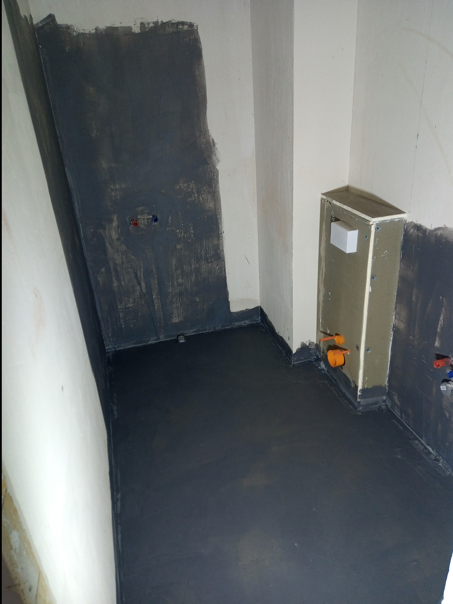 Łazienka w stanie deweloperskim: wykonana hydroizolacja ścian i podłogi w kolorze grafitowym, widoczna zabudowa stelaża WC z instalacją hydrauliczną.